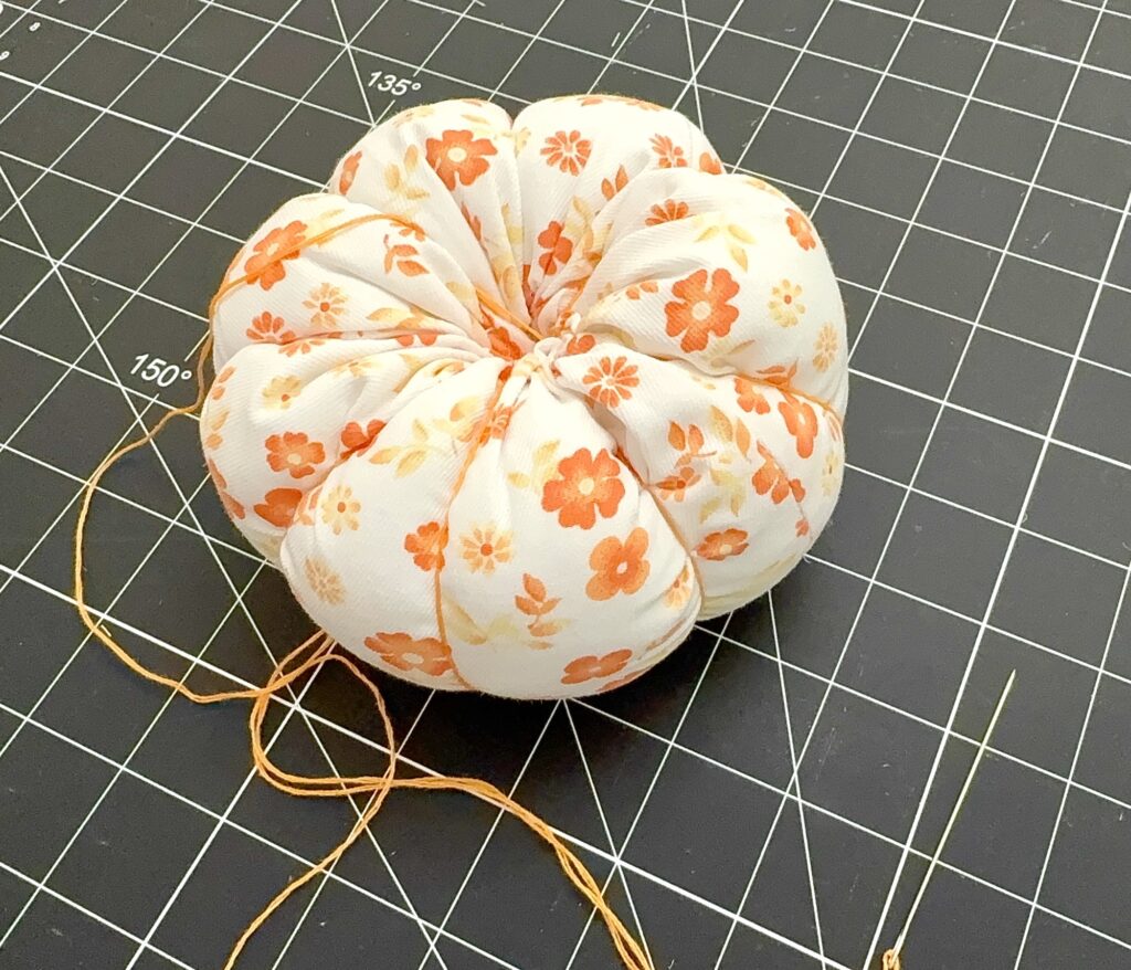 A round fabric pincushion with orange flowers, yellow thread, and a sewing needle rests on a black cutting mat with white measurement lines.