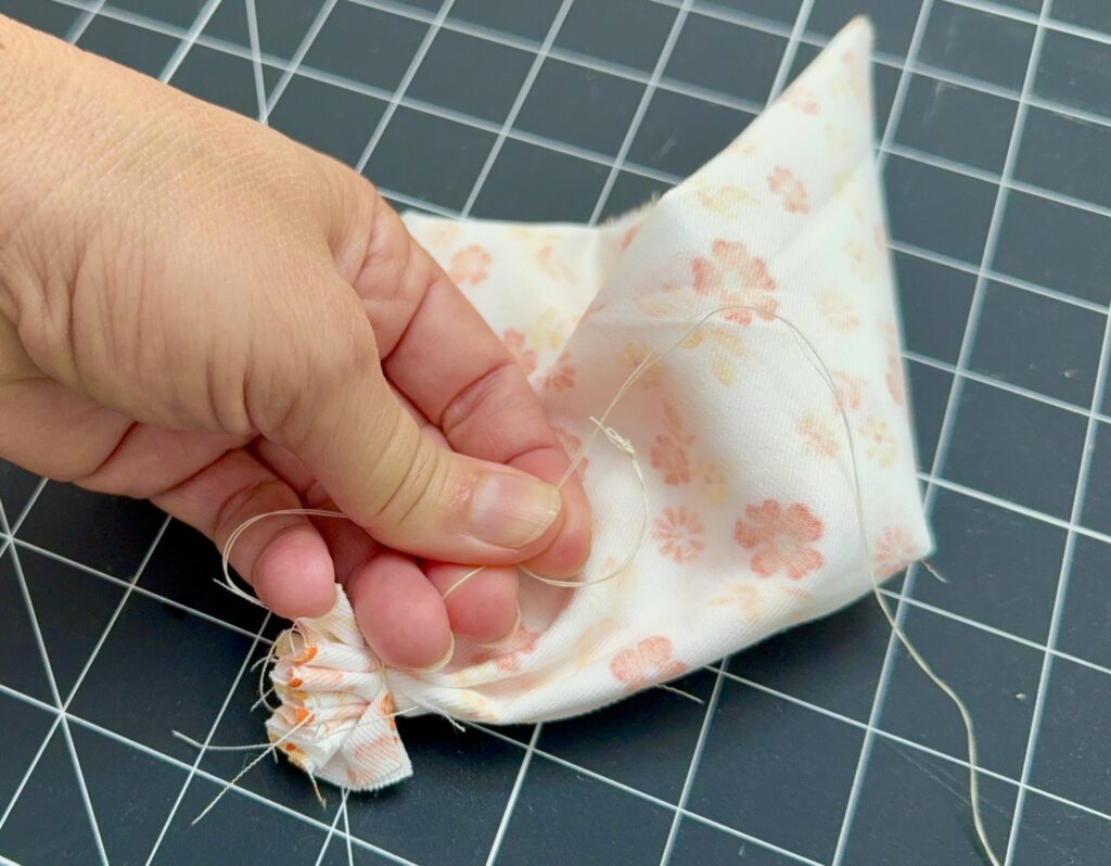 A hand is sewing closed the gathered edge of a white fabric with pink floral patterns on a grid cutting mat.