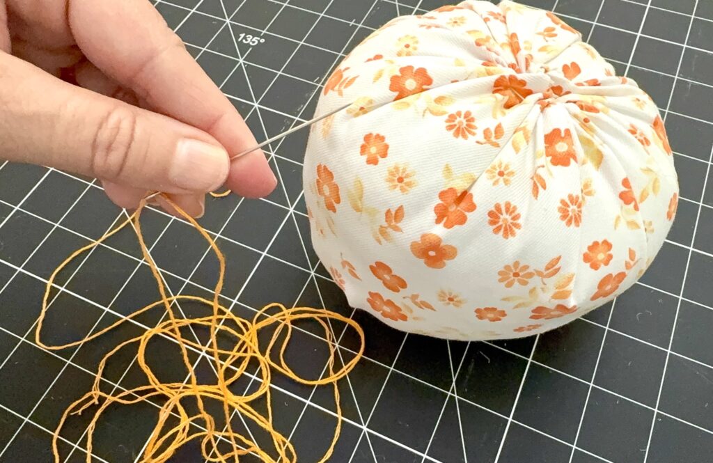 A hand is sewing closed a fabric ball with orange floral print using a needle and orange thread on a black grid cutting mat.
