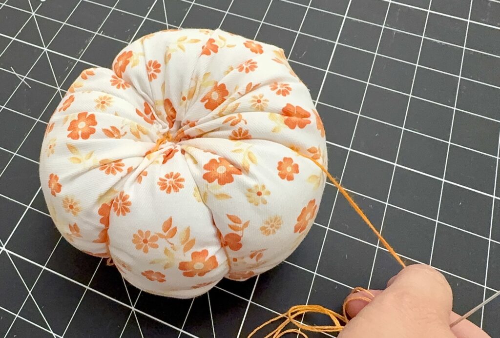 A hand holds orange thread while sewing an orange and white floral fabric pincushion on a black grid cutting mat.
