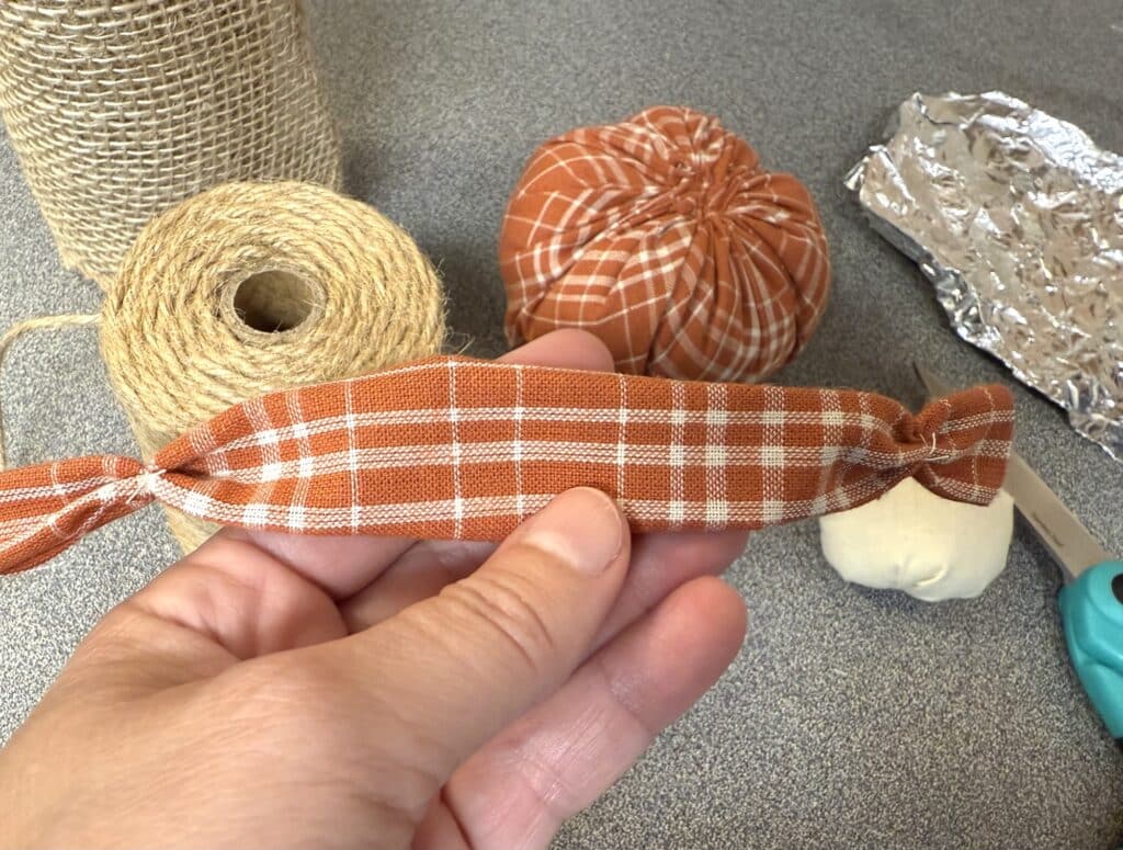 A hand holds a small piece of orange plaid fabric tied at both ends, with twine, burlap, fabric pumpkins, foil, and scissors on a gray surface in the background.