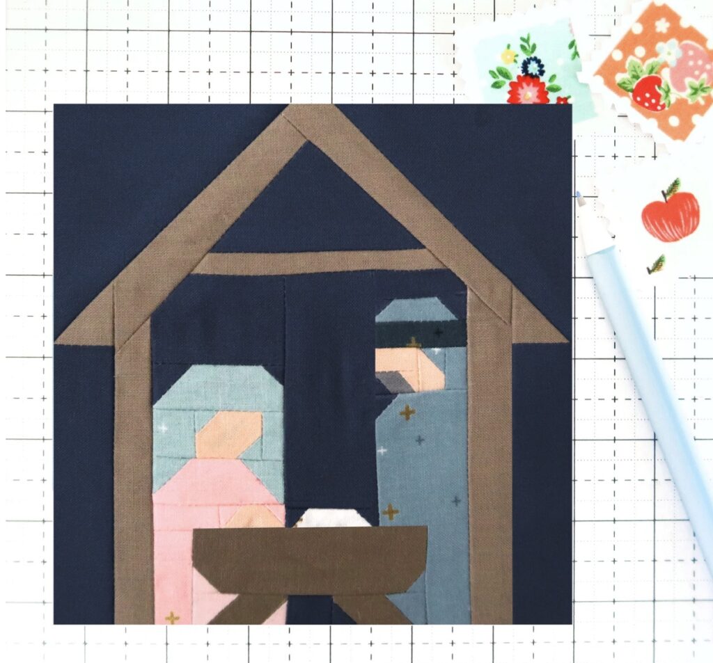 A Christmas quilt block depicts a nativity scene with stylized fabric figures of Mary, Joseph, and baby Jesus in a manger under a stable roof on a grid background.