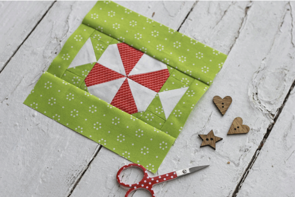 A quilt block with a red and white pinwheel pattern and green floral border is on a white wooden surface, next to red scissors and wooden heart and star buttons.