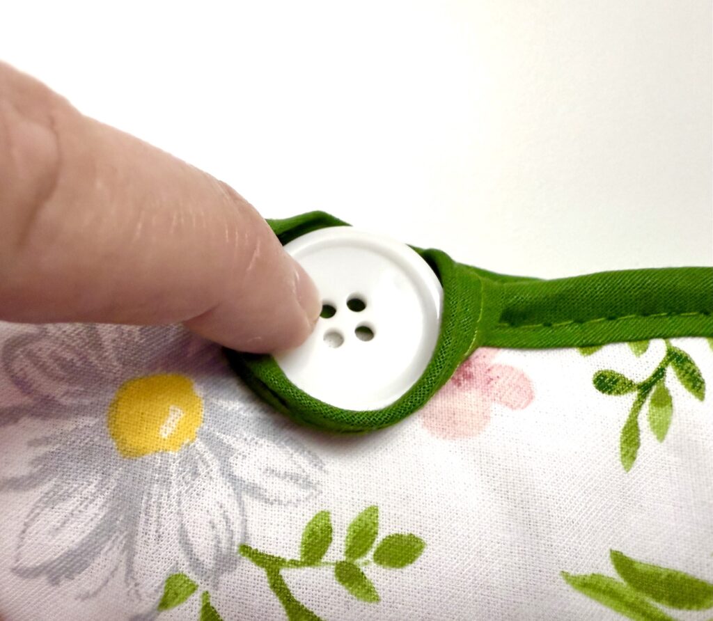 A hand is pressing a white four-hole button attached to green trim on a floral-patterned pot holder.