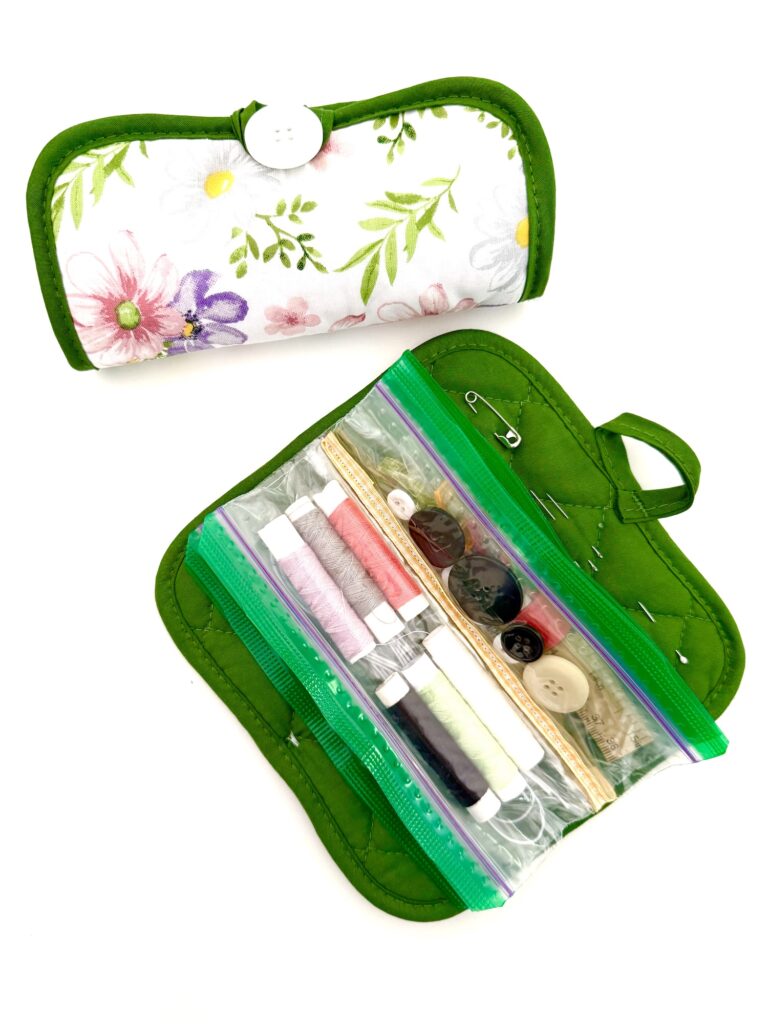 A compact sewing kit with a floral-patterned fabric cover and green trim, opened to reveal thread spools, buttons, needles, pins, a pot holder accent, and a handy zipper compartment.