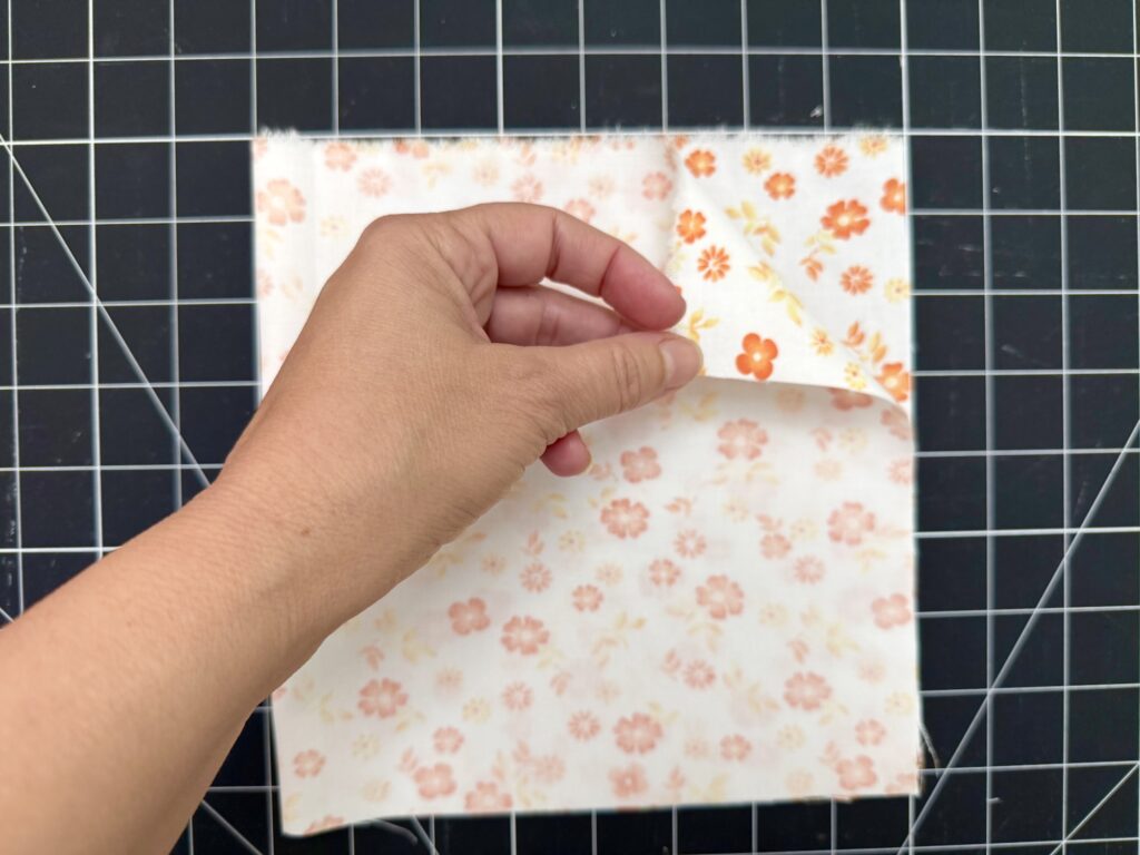 A hand lifts the top corner of a square piece of floral fabric placed on a black grid cutting mat.