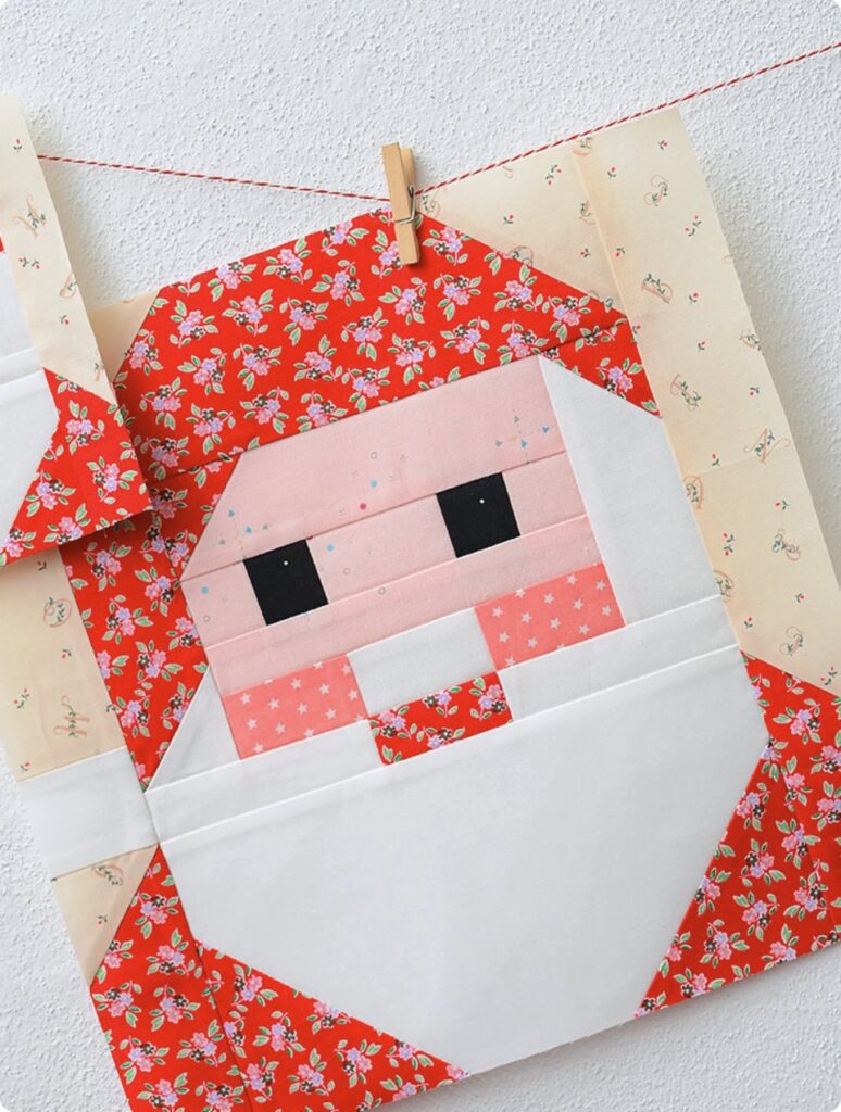 A quilt block features a geometric Santa Claus face with a red and white patterned border, hanging from a clothespin against a light wall.