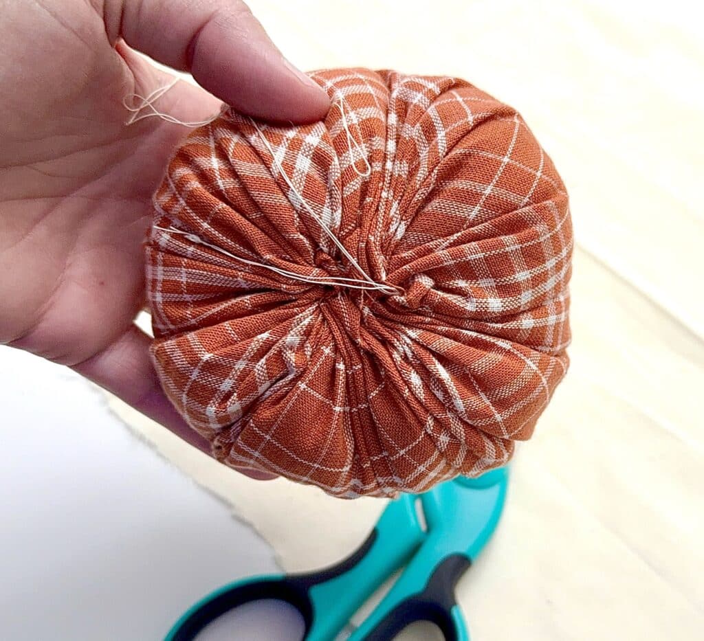 A hand holds a round, gathered piece of orange plaid fabric; blue scissors rest on the surface below.