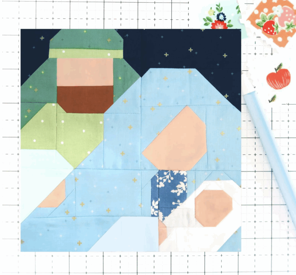 Patchwork quilt block depicting a stylized nativity scene with geometric shapes for figures, set on a grid background with sewing tools nearby—a charming addition to your Christmas quilt blocks collection.