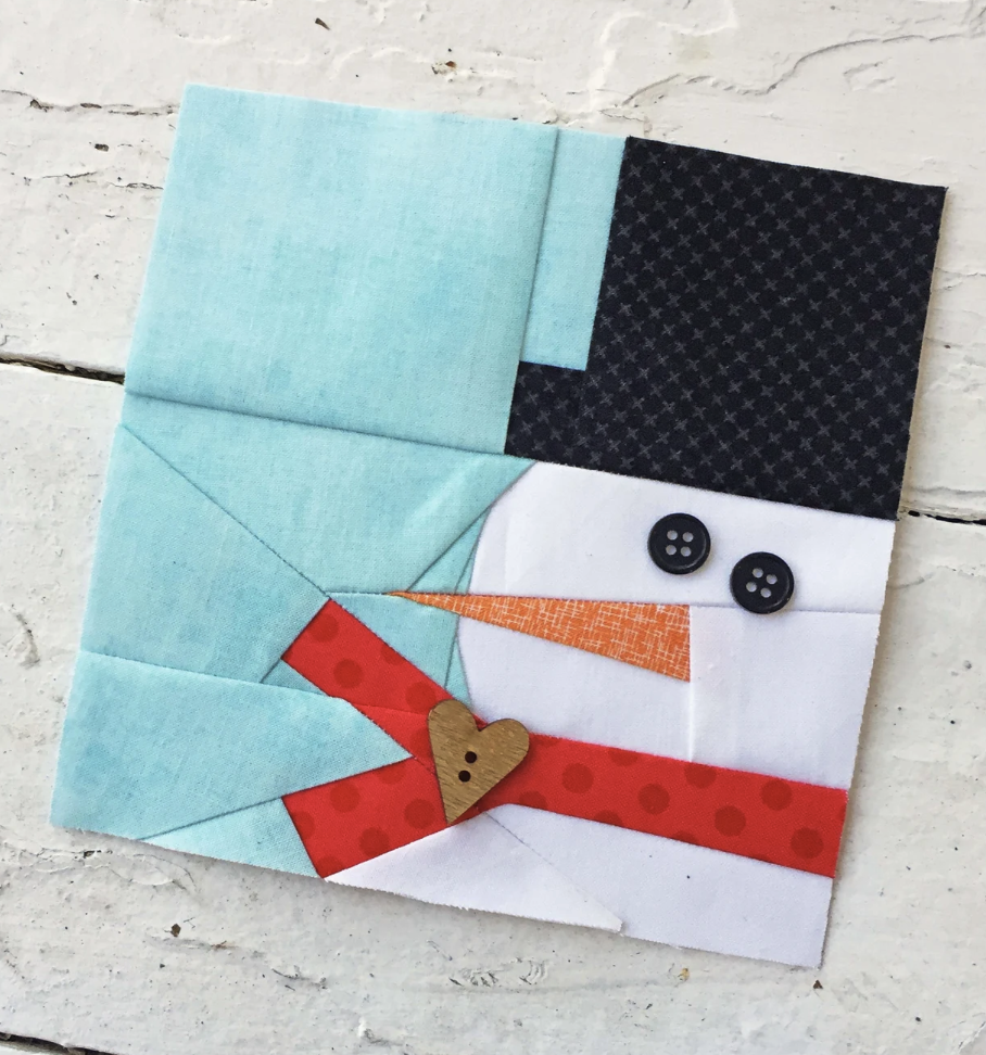 A Christmas quilt block featuring a snowman with a blue hat, red scarf, orange nose, button eyes, and a wooden heart button on a white wooden surface.