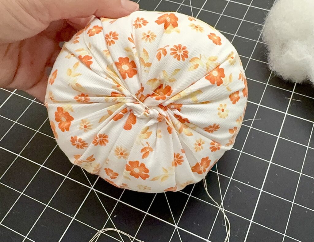 A hand holds a round, fabric item with orange floral print, gathered at the center; white stuffing and thread are visible on a black grid surface.