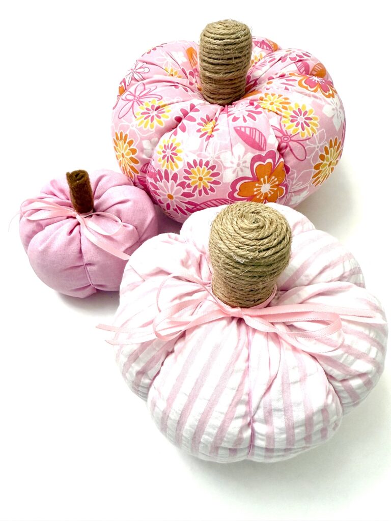 Three fabric pumpkins in pink and floral patterns with twine-wrapped stems are displayed on a white background.