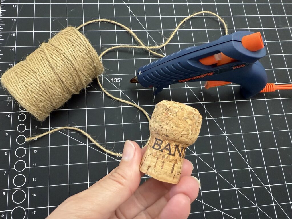 A hand holds a champagne cork labeled "BAN" in front of a glue gun, twine, and a cutting mat with measurement markings.