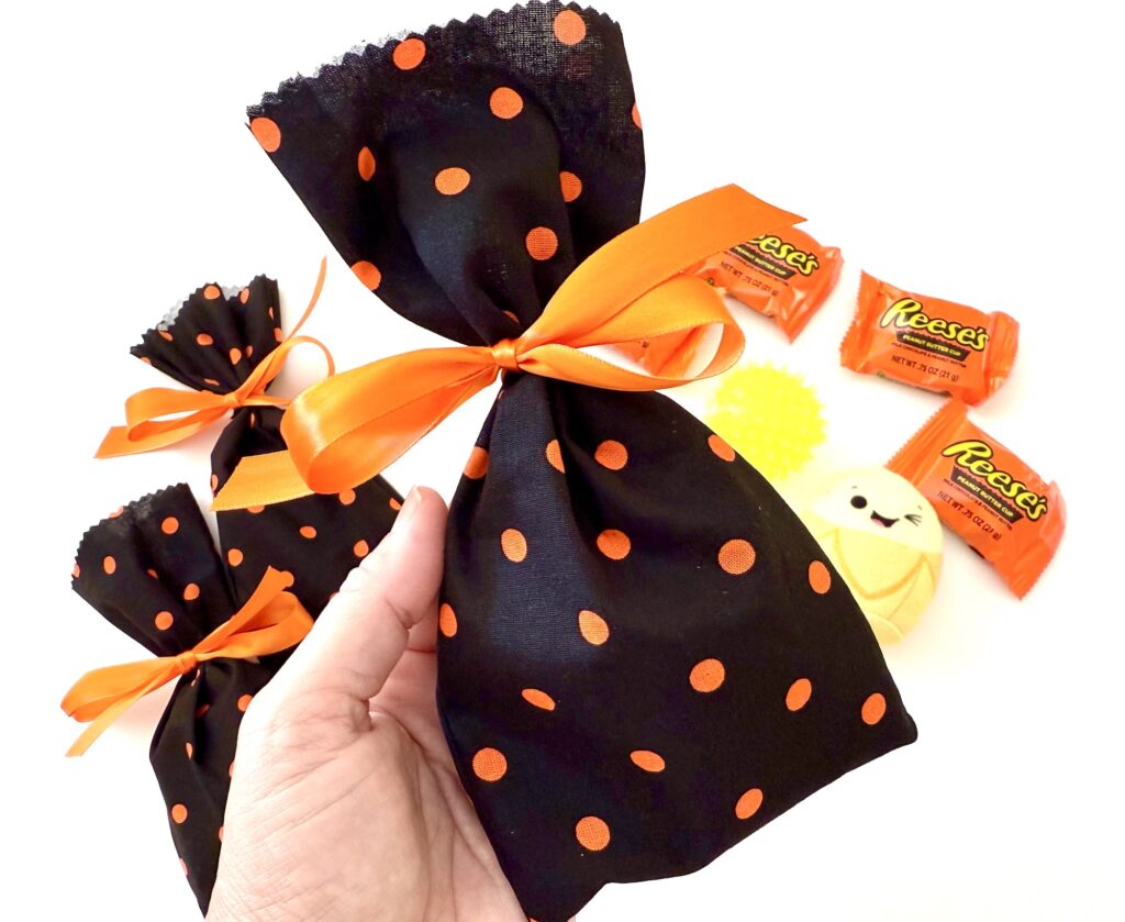 A hand holds a black fabric gift bag with orange polka dots and an orange ribbon. More similar bags and Halloween treats are in the background.
