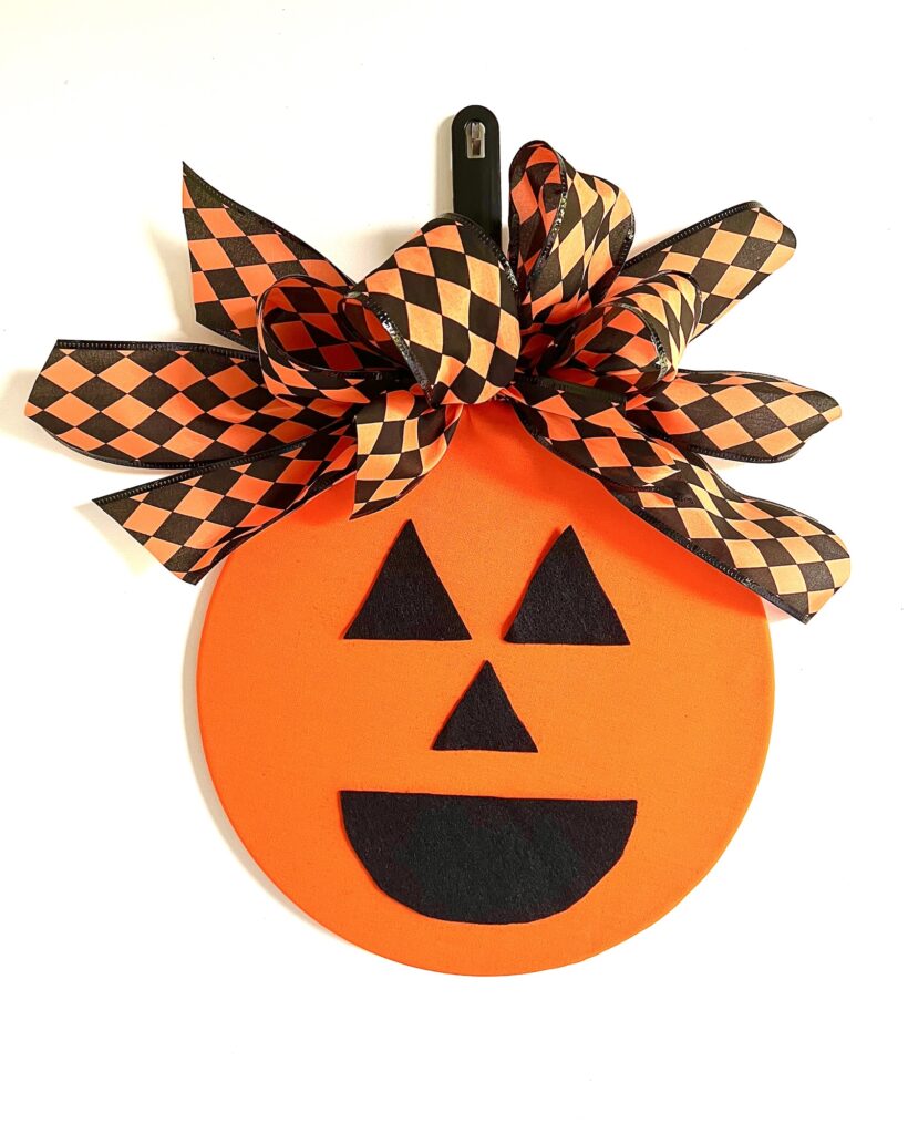 An orange round Halloween wreath resembling a jack-o'-lantern face with black eyes, nose, and mouth, topped with a large patterned bow in black and orange diamond shapes.