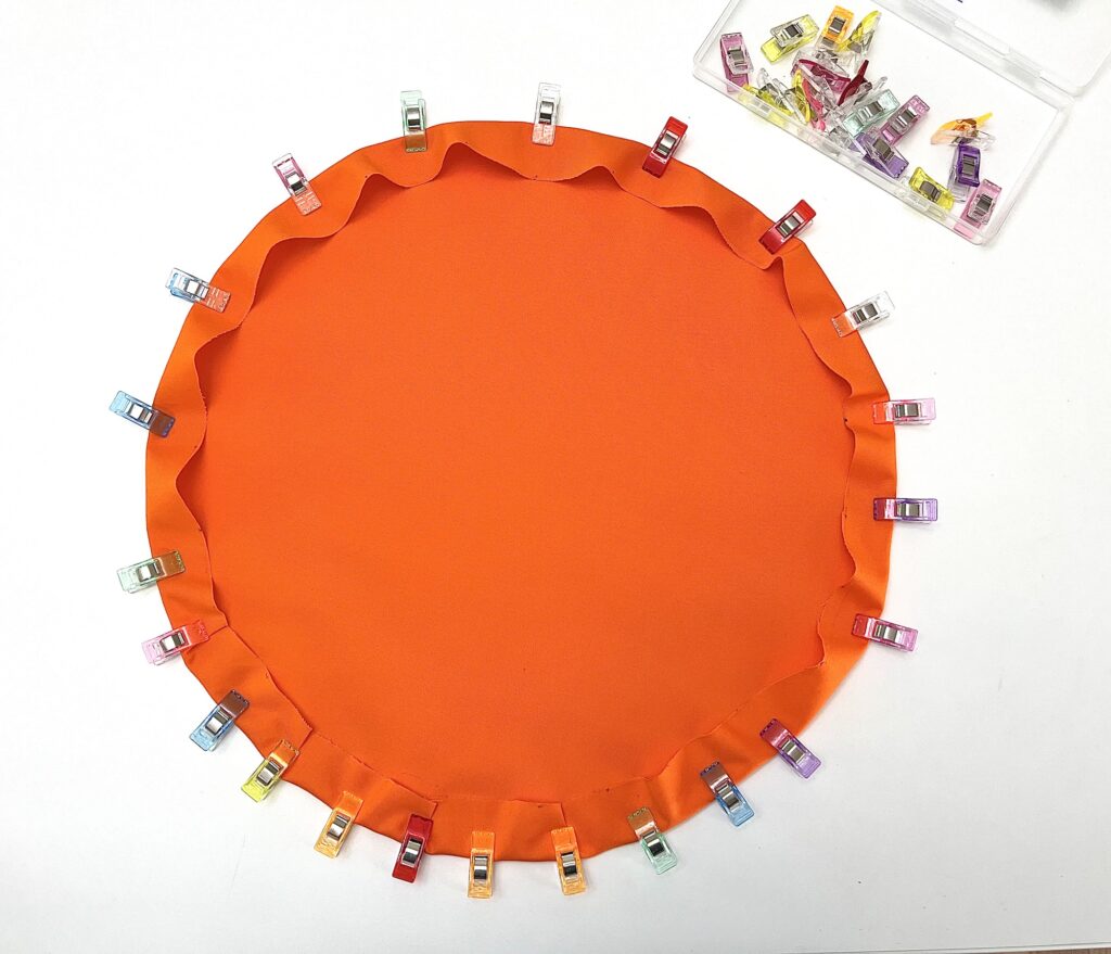 A round piece of orange fabric with its edges folded and secured by colorful sewing clips, with a box of additional clips nearby on a white surface.