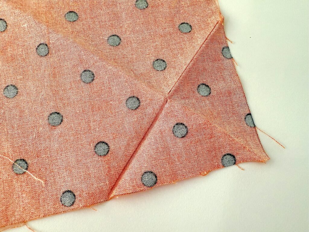 A piece of light orange fabric with black polka dots is folded diagonally on a white surface, resembling an over the collar dog bandana, with visible stitches and raw edges.