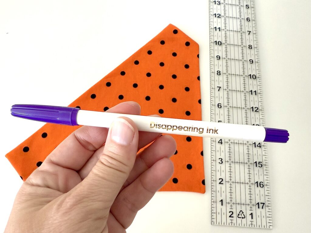 A hand holding a purple disappearing ink pen above an orange polka dot over the collar dog bandana and a transparent ruler.