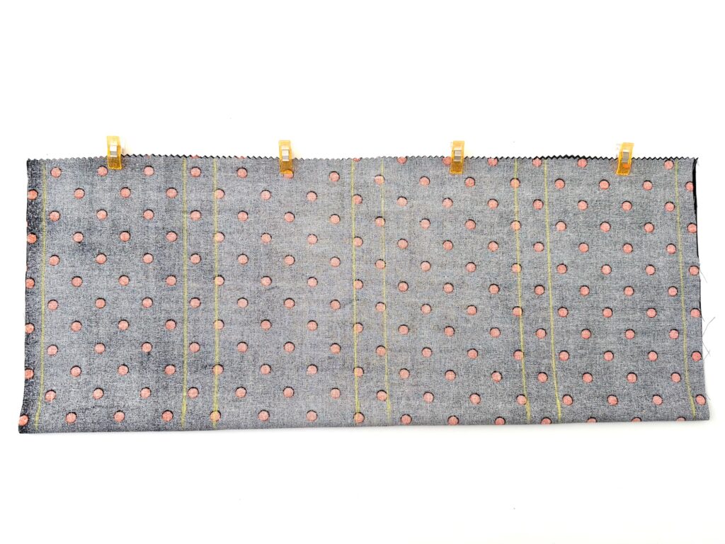 A rectangular piece of fabric with a pink polka dot pattern is clipped at the top with five yellow clips and has four vertical lines of yellow stitching.