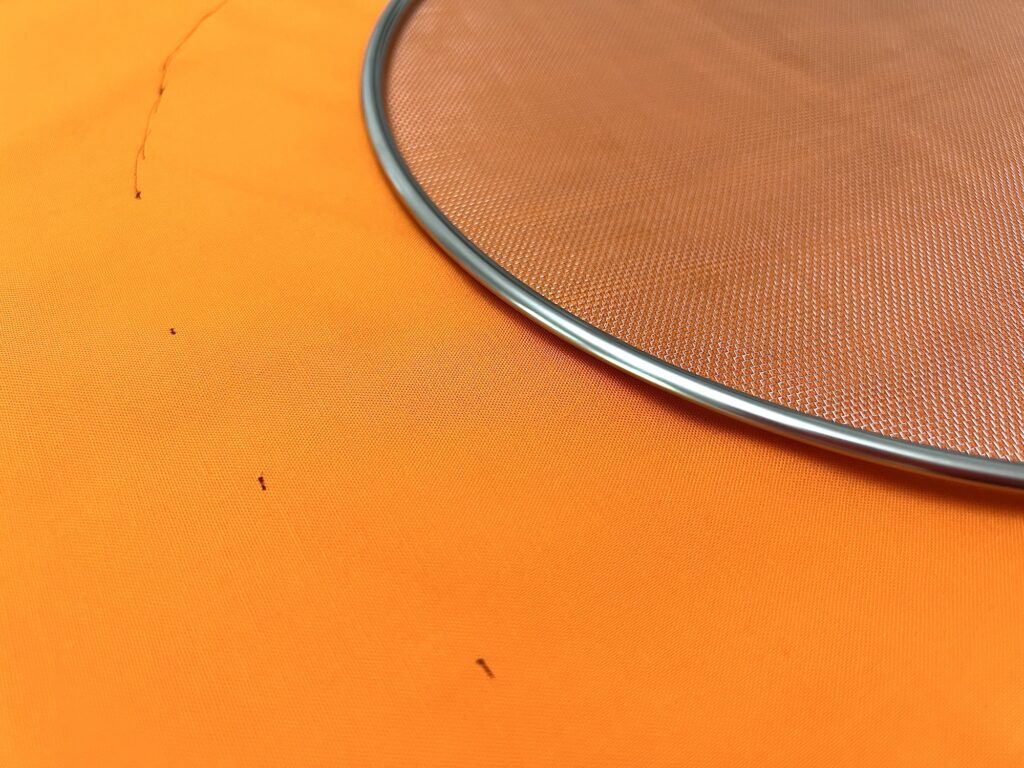 A round metal mesh strainer is partially visible on a bright orange fabric background with a few small black threads.