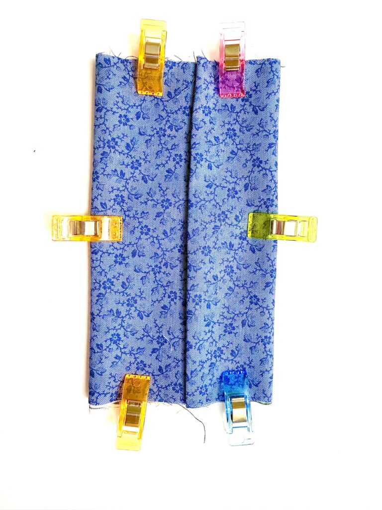 A piece of blue floral fabric is folded and secured with six colorful plastic clips on a white background.