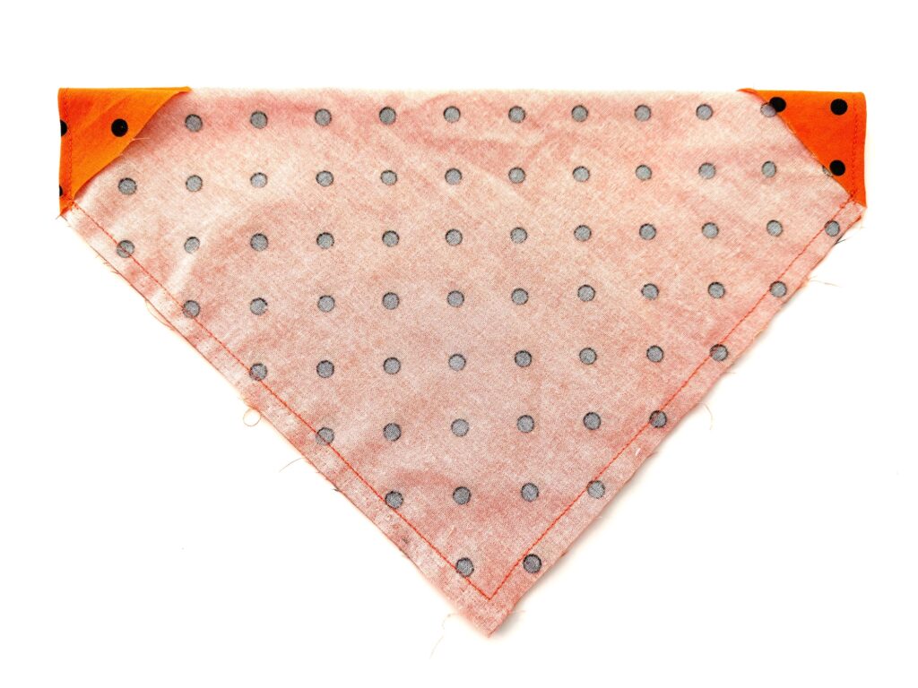A triangular folded light pink over the collar dog bandana with black polka dots and orange corners is displayed on a white background.