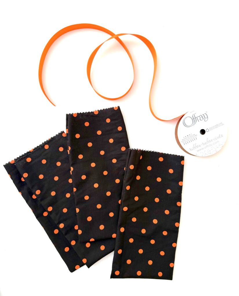 Three rectangular pieces of black fabric with orange polka dots and a spool of orange ribbon are arranged on a white background.