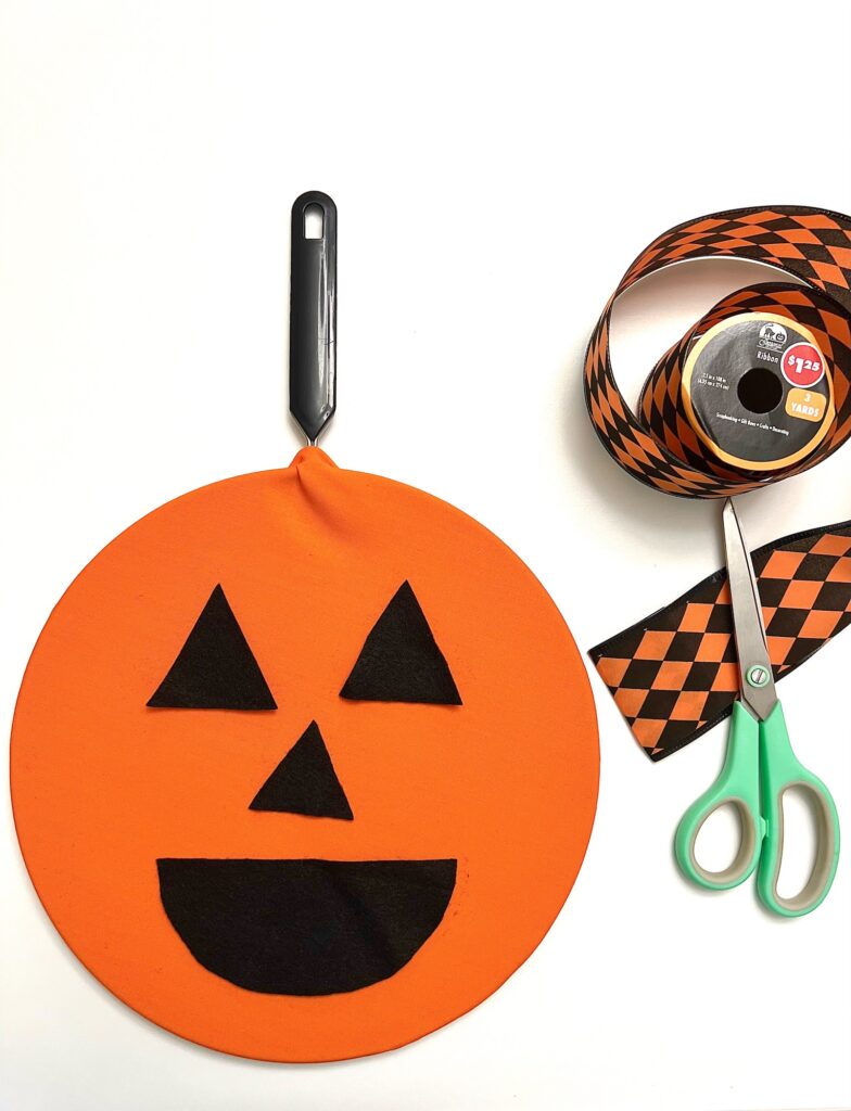 A round orange spatula cover with a jack-o'-lantern face, a roll of orange and black checkered ribbon, and green-handled scissors on a white surface—perfect supplies for crafting a festive Halloween wreath.