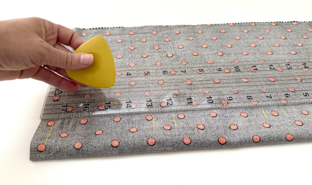 A hand marks fabric with a yellow chalk while measuring with a clear ruler; the fabric has a gray background with orange dots.