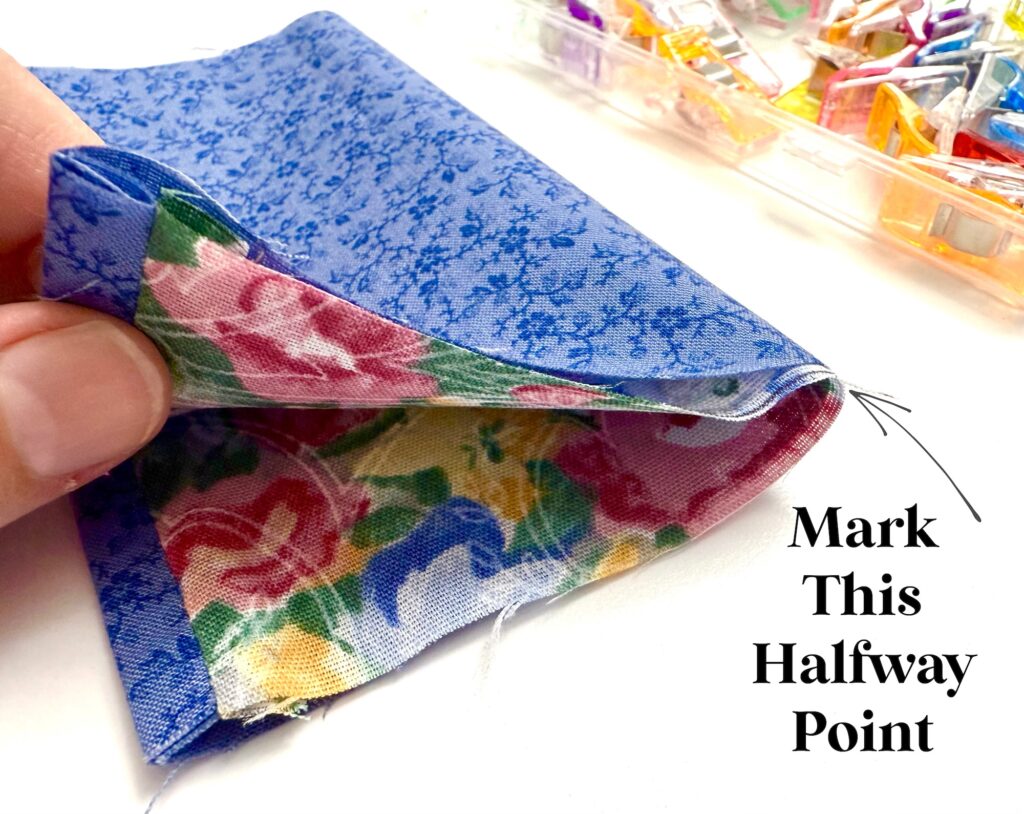 A hand holds folded blue floral fabric with a colorful patterned lining, and text reads "Mark This Halfway Point." Fabric clips are visible in the background.