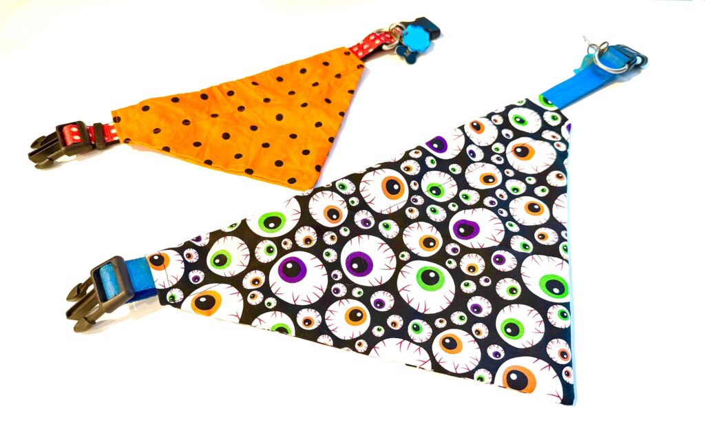 Two over the collar dog bandanas: one orange with black polka dots and one black with a colorful eyeball pattern, both laid flat on a white background.