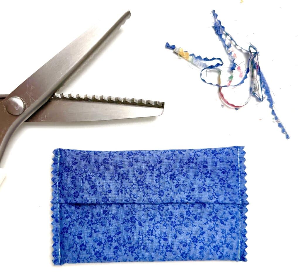 A pair of pinking shears, fabric scraps, and a blue floral tissue holder with zigzag-edged seams are arranged on a white surface.
