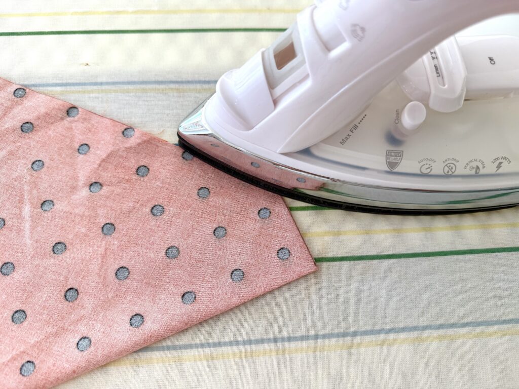 A white iron is pressing a pink polka dot over the collar dog bandana on an ironing board with a striped cover.