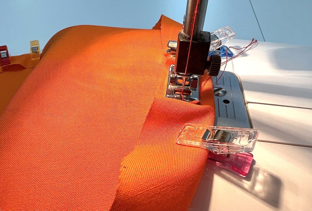 Close-up of a sewing machine stitching bright orange fabric, held in place by colorful plastic clips.