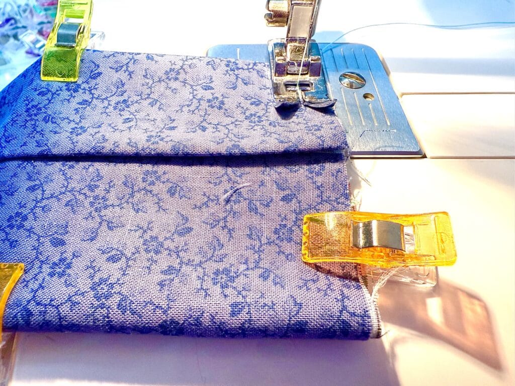 Close-up of a sewing machine stitching blue patterned fabric, secured with yellow and orange clips.