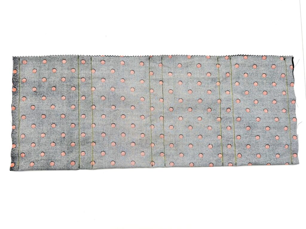 A rectangular piece of gray fabric with a repeating pink polka dot pattern, featuring vertical fold lines and zigzag-cut edges, is displayed on a white background.
