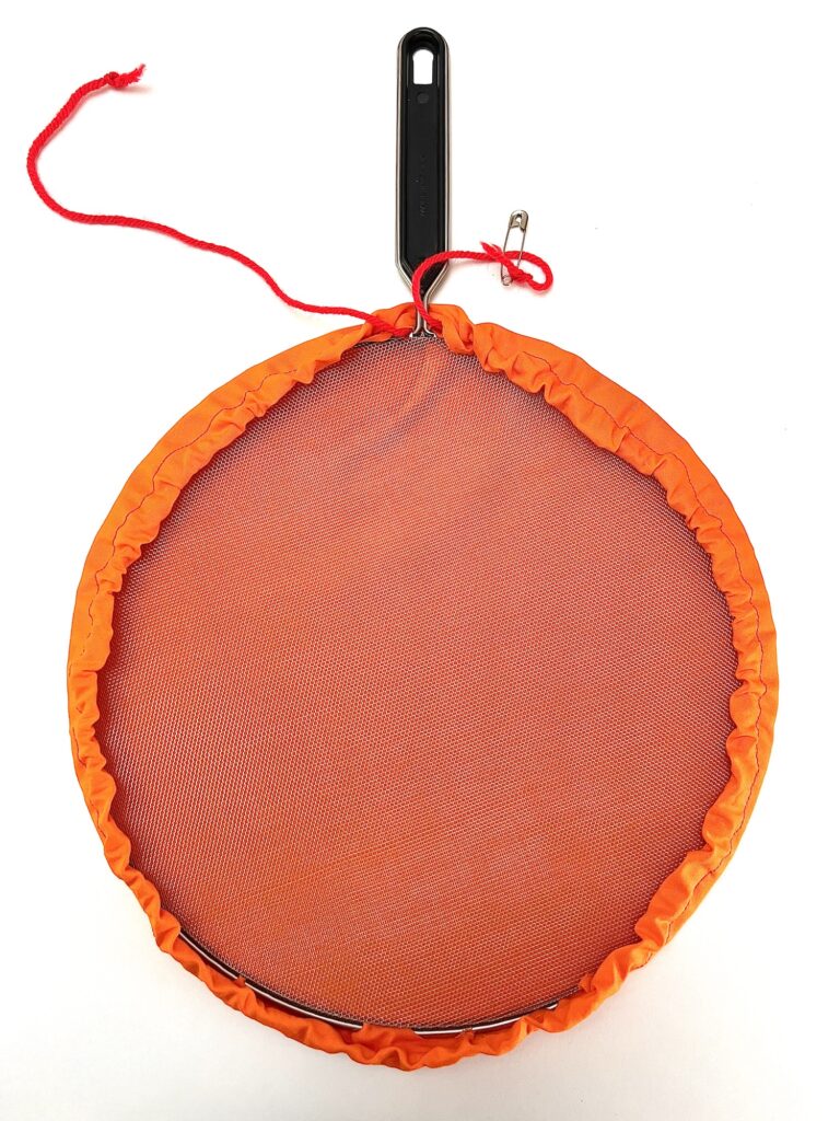 A round, mesh splatter screen with an orange fabric edge and a black handle, lying flat on a white surface. A red string is attached to the handle, making it perfect as the base for a DIY Halloween wreath.