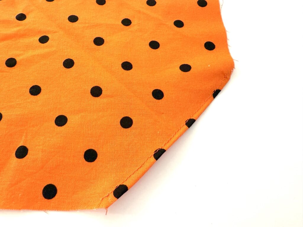 An orange over the collar dog bandana featuring a pattern of evenly spaced black polka dots, shown with one stitched and slightly frayed edge.