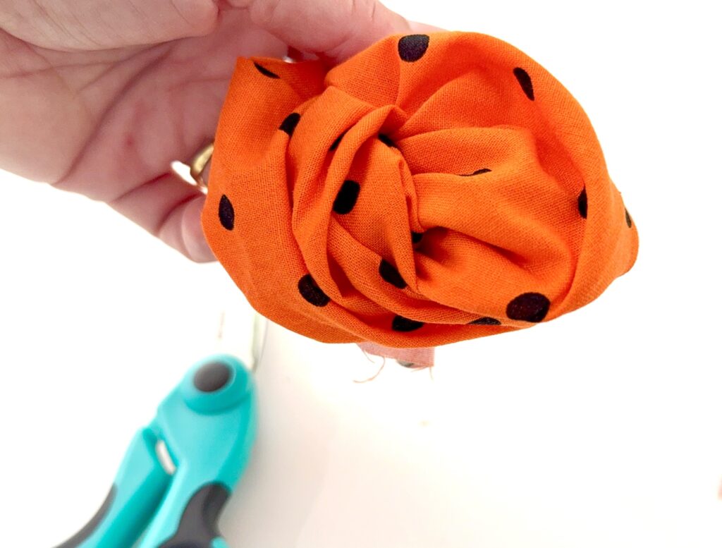 A hand holds an orange fabric flower with black polka dots, perfect for decorating an over the collar dog bandana; blue-handled scissors are visible in the background.