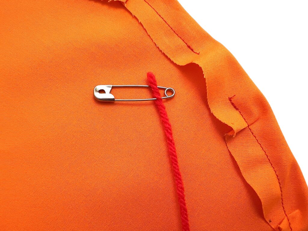 A safety pin attaches a red yarn to an orange fabric with unfinished edges and visible seams.