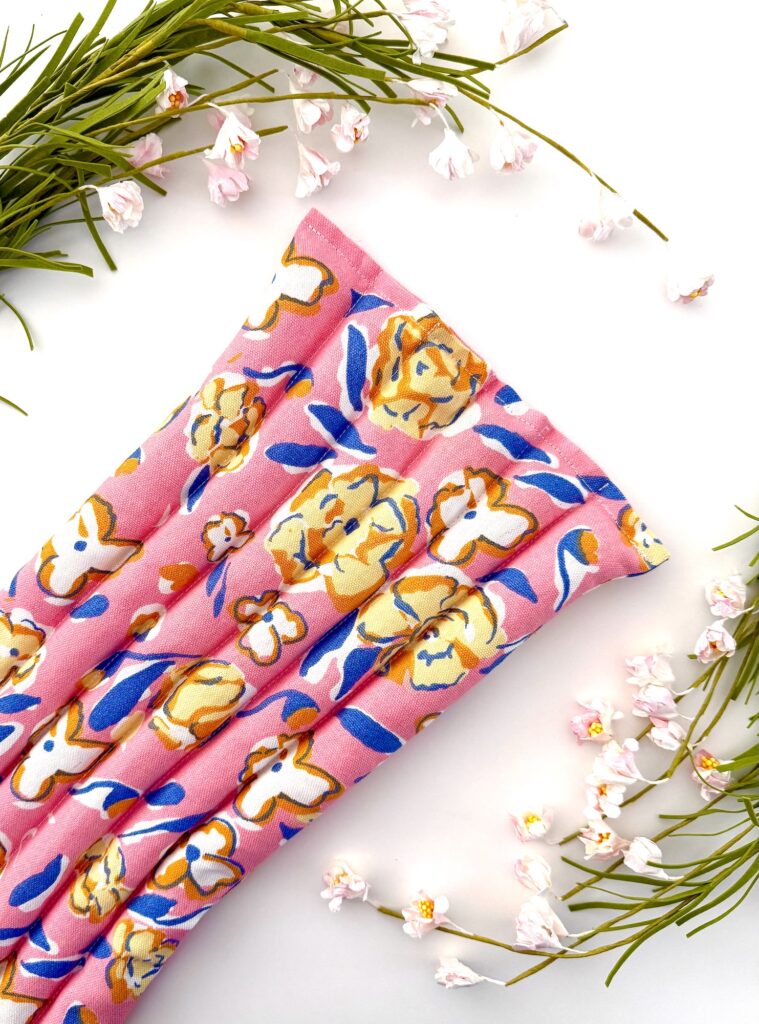 A pink fabric trivet with a yellow and white floral pattern is placed diagonally among green sprigs and small pink flowers on a white background.