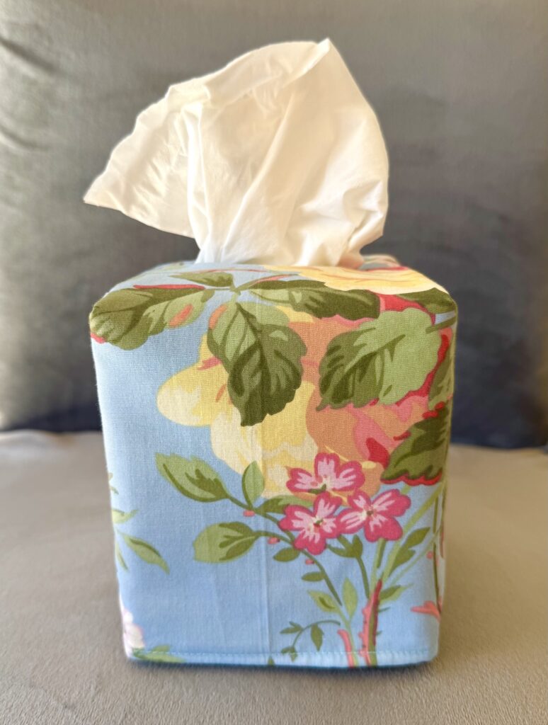 A tissue box cover in blue fabric with a floral pattern neatly wraps a box of tissues, with one tissue partially pulled out.