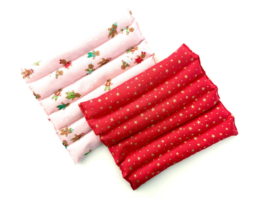 Two rectangular fabric heat pads, one pink with gingerbread designs and one red with gold stars, are placed side by side on a white background.