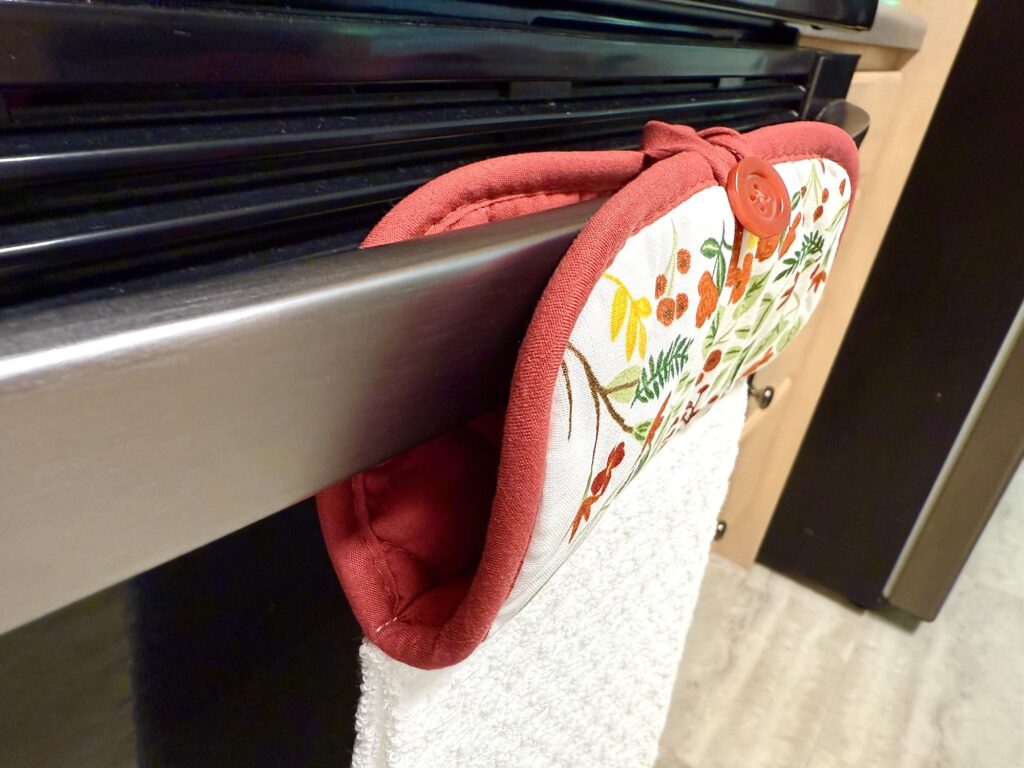 A red and white pot holder with a botanical pattern hangs on an oven handle, with a white kitchen towel draped underneath it.