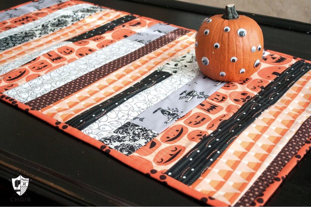 A Halloween-themed quilted table runner with orange, black, and white strips, topped with a small pumpkin decorated with googly eyes.