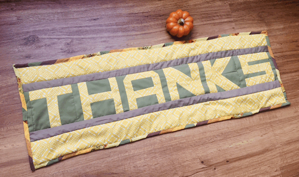 A quilted table runner with the word "THANKS" in large letters lies on a wooden floor next to a small pumpkin.