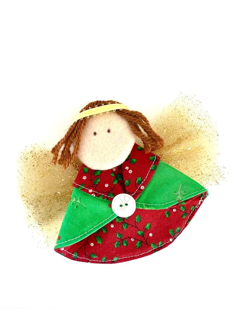 Handmade fabric angel ornament with brown yarn hair, a green and red dress, a white button, and gold wings, set against a white background.