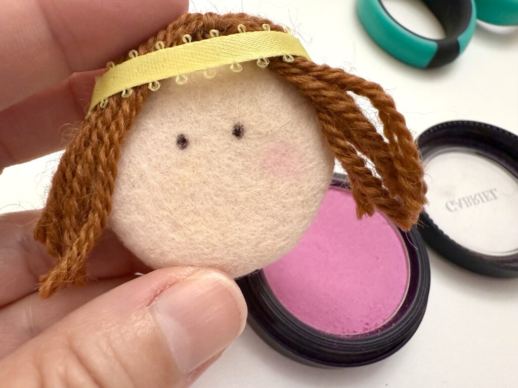 A hand holds a round doll face made of fabric with brown yarn hair, next to an open container of pink blush makeup.