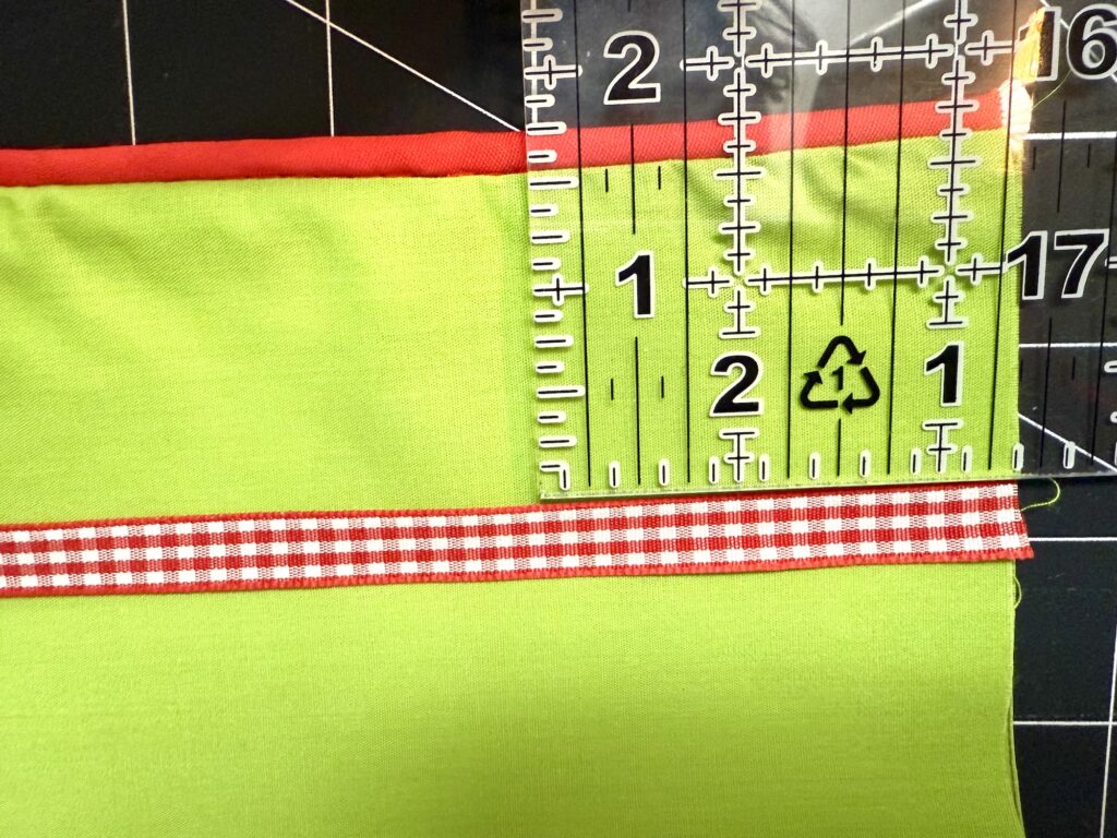 A clear ruler measures green fabric for an advent tree, with a red gingham ribbon and a red trim on top, placed on a cutting mat with grid lines and measurement markings.