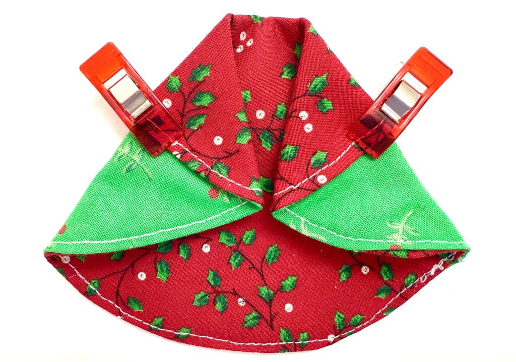 A piece of red and green fabric with a holly pattern is folded and held in place by two red sewing clips on a white background.